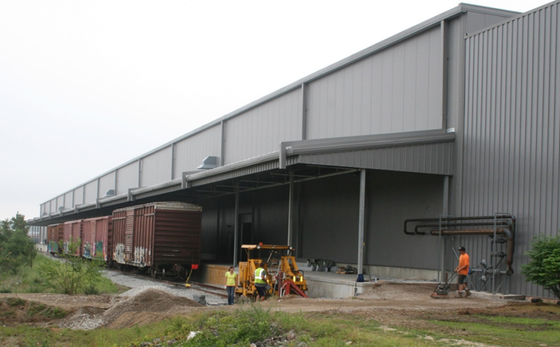 FIGURE 2. New Rail Siding Adjacent Expanded Warehouse
This image shows four railroad boxcars positioned next to a loading dock along the side of a modern warehouse.
FIGURE 2. New Rail Siding Adjacent Expanded Warehouse
This image shows four railroad boxcars positioned next to a loading dock along the side of a modern warehouse.